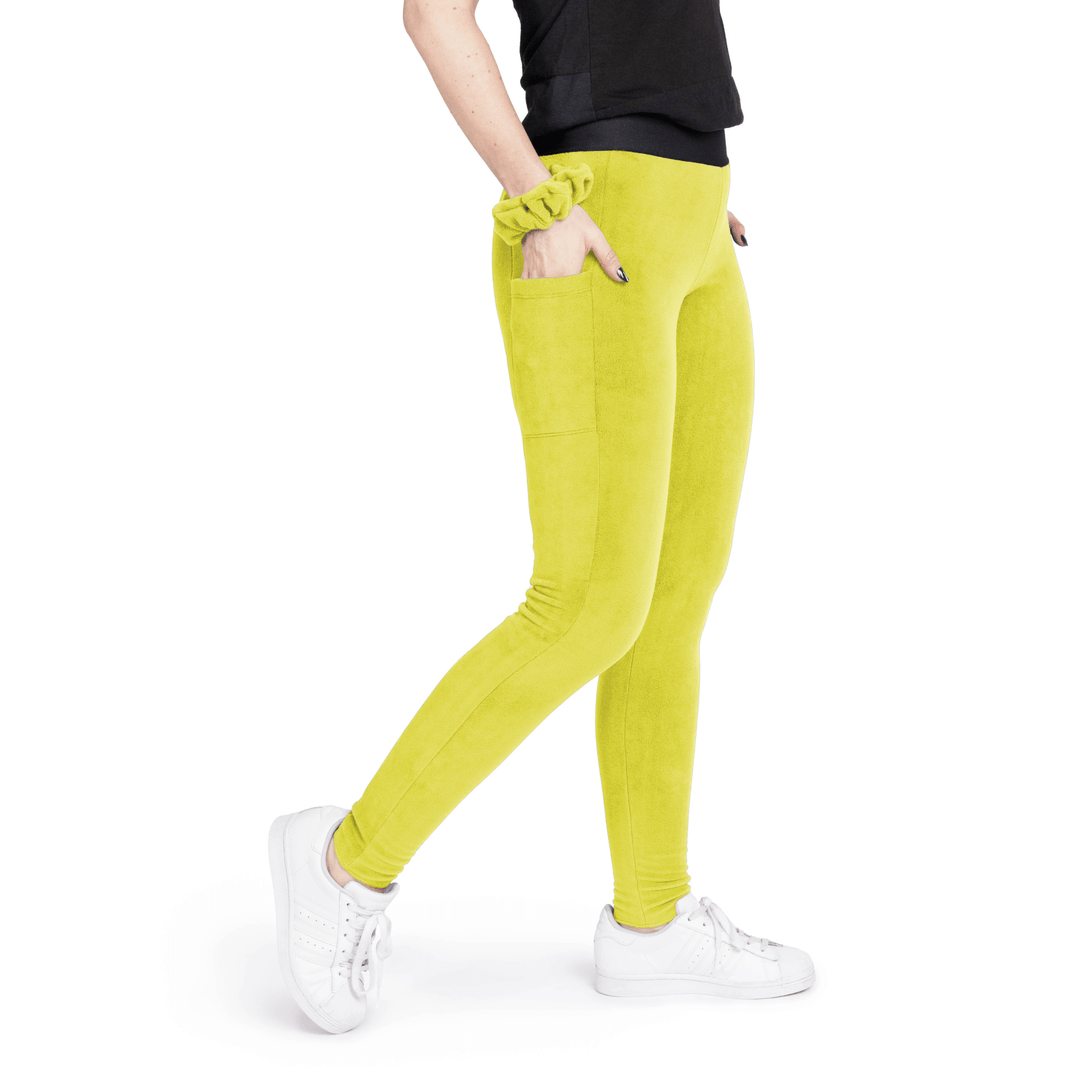 A woman stands wearing a black t shirt and avocado green fleece leggings with a matching scrunchie on their wrist in front of a white background