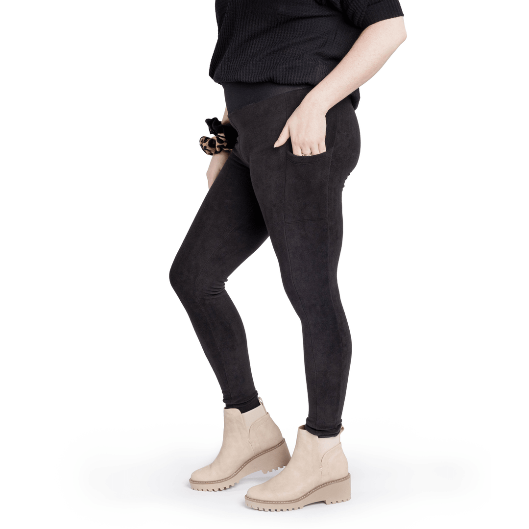 A woman stands wearing a black t shirt and black fleece leggings with a matching scrunchie on their wrist in front of a white background