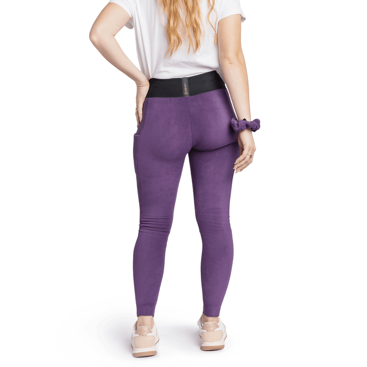 A woman stands wearing a black t shirt and huckleberry purple fleece leggings with a huckleberry purple scrunchie on their wrist in front of a white background