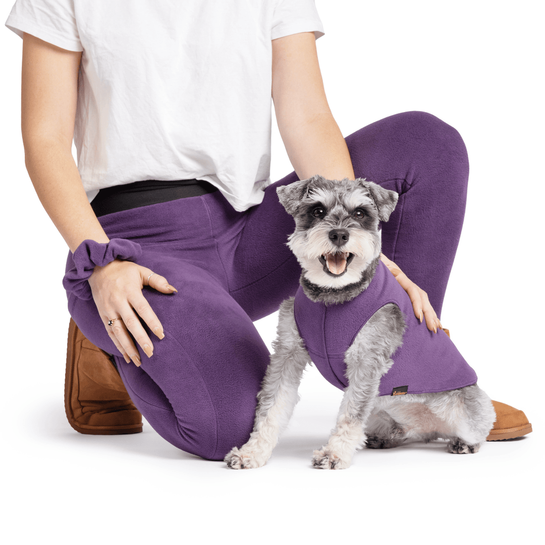 A woman kneels wearing huckleberry purple fleece leggings with a huckleberry scrunchie on her wrist. In front of her is a mini schnauzer in a huckleberry purple stretch fleece. They are in front of a white background