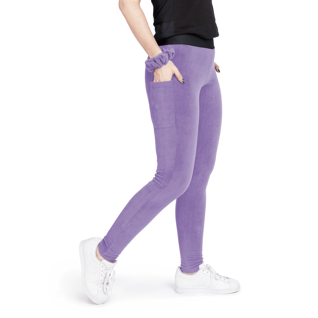 A woman stands wearing a black t shirt and lavender fleece leggings with a lavender scrunchie on their wrist in front of a white background