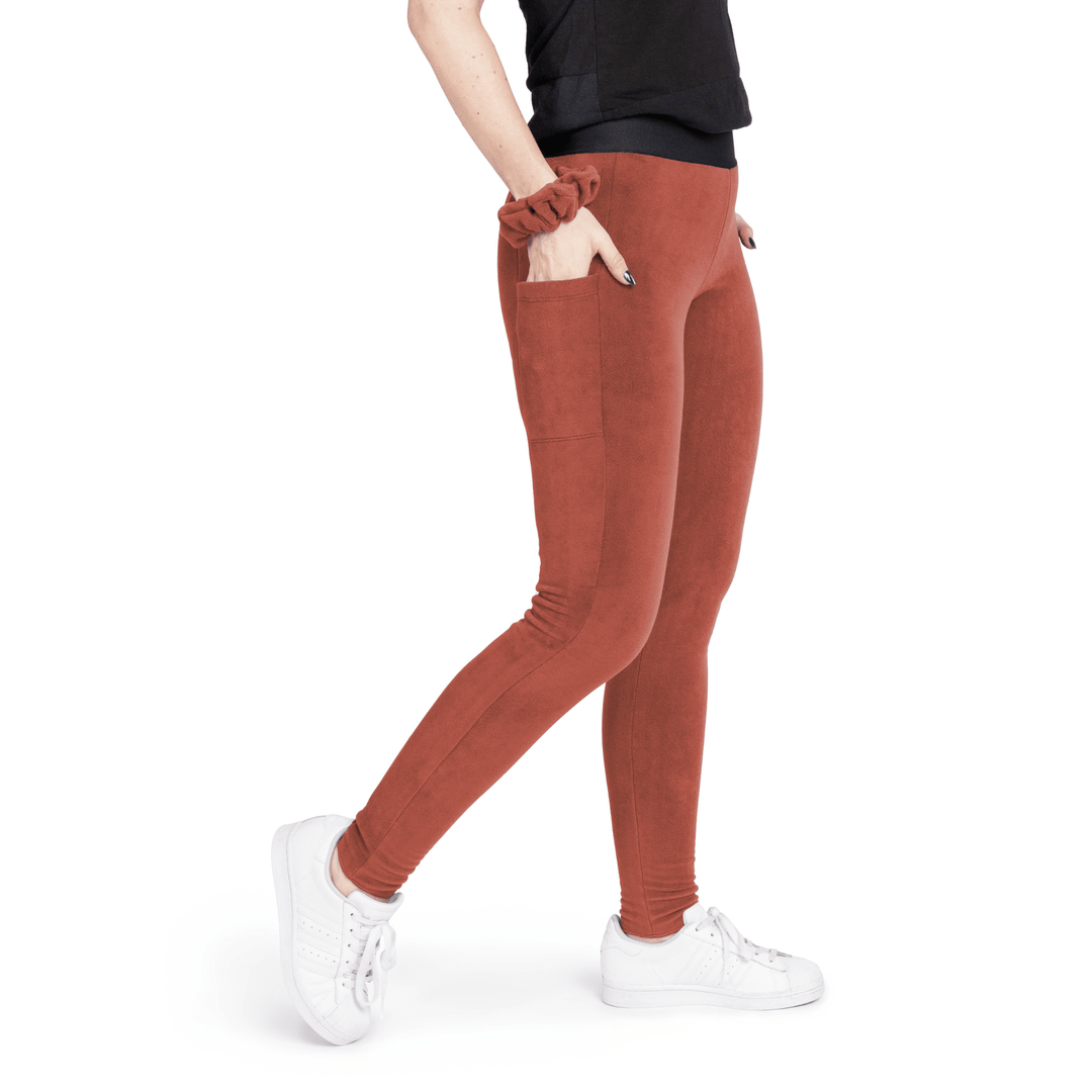 A woman stands wearing a black t shirt and paprika orange fleece leggings with a paprika orange scrunchie on their wrist in front of a white background