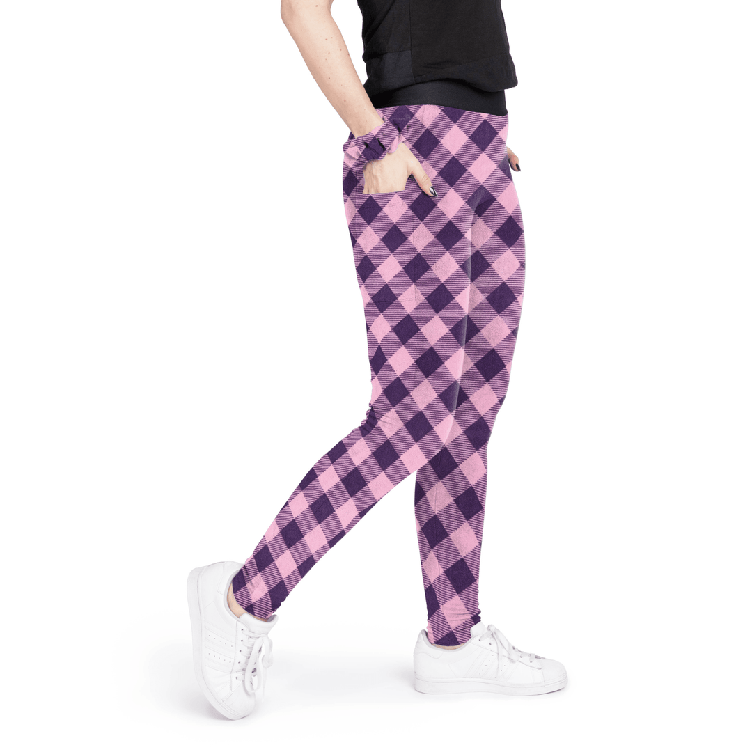 A woman stands wearing a black t shirt and rose pink and huckleberry purple buffalo check fleece leggings with a matching scrunchie on their wrist in front of a white background